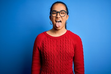 Young beautiful asian woman wearing casual sweater and glasses over blue background sticking tongue out happy with funny expression. Emotion concept.