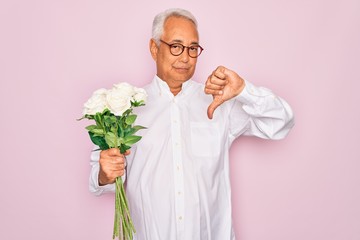 Middle age senior grey-haired man holding romantic bouquet of white roses over pink background with angry face, negative sign showing dislike with thumbs down, rejection concept