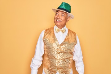 Middle age senior grey-haired man wearing Brazilian carnival custome over yellow background looking away to side with smile on face, natural expression. Laughing confident.