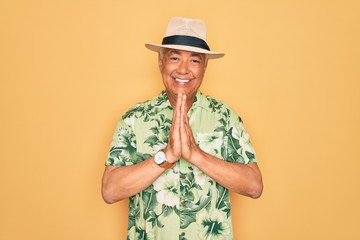 Middle age senior grey-haired man wearing summer hat and floral shirt on beach vacation praying with hands together asking for forgiveness smiling confident.