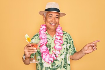 Middle age senior grey-haired man wearing summer hat and hawaiian lei drinking a cocktail very...