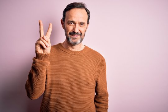 Middle age hoary man wearing casual brown sweater standing over isolated pink background smiling with happy face winking at the camera doing victory sign. Number two.