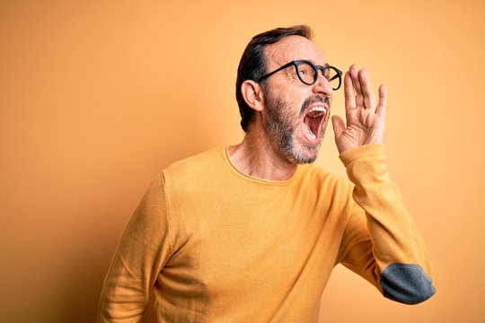Middle age hoary man wearing casual sweater and glasses over isolated yellow background shouting and screaming loud to side with hand on mouth. Communication concept.
