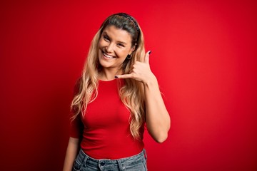 Young beautiful blonde woman wearing casual t-shirt standing over isolated red background smiling doing phone gesture with hand and fingers like talking on the telephone. Communicating concepts.