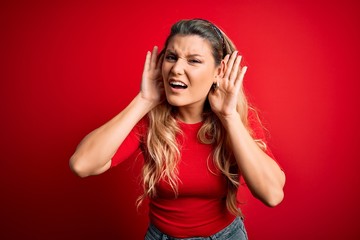 Young beautiful blonde woman wearing casual t-shirt standing over isolated red background Trying to hear both hands on ear gesture, curious for gossip. Hearing problem, deaf