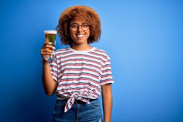 Young beautiful African American afro woman with curly hair on vacation drinking glass of beer with a happy face standing and smiling with a confident smile showing teeth