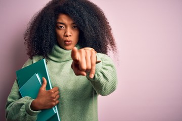 Young african american student woman with afro hair reading books over pink background pointing...