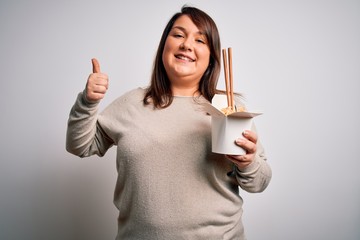 Beautiful plus size woman eating asian noodles food from box over isolated background happy with big smile doing ok sign, thumb up with fingers, excellent sign