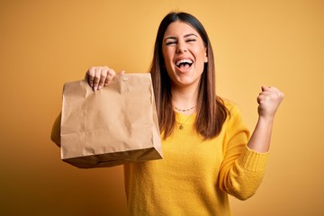 Young beautiful woman holding take away paper bag from delivery over yellow background screaming...