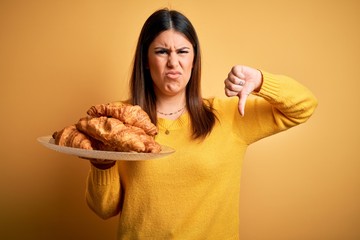 Young beautiful woman holding french croissant pastry over yellow background with angry face, negative sign showing dislike with thumbs down, rejection concept