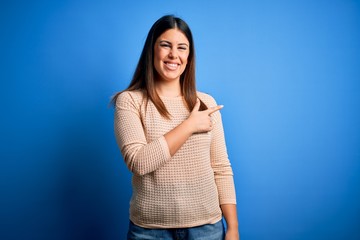 Young beautiful woman wearing casual sweater over blue background cheerful with a smile of face pointing with hand and finger up to the side with happy and natural expression on face