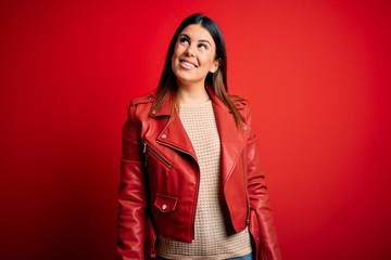 Young beautiful woman wearing red leather jacket over isolated background looking away to side with smile on face, natural expression. Laughing confident.