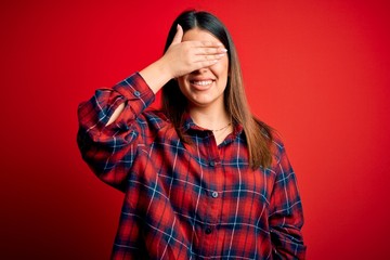 Young beautiful woman wearing casual shirt over red background smiling and laughing with hand on face covering eyes for surprise. Blind concept.