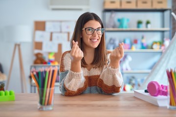 Young beautiful teacher woman wearing sweater and glasses sitting on desk at kindergarten doing money gesture with hands, asking for salary payment, millionaire business