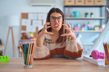 Young beautiful teacher woman wearing sweater and glasses sitting on desk at kindergarten Shouting...
