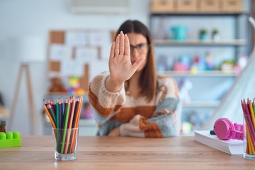 Young beautiful teacher woman wearing sweater and glasses sitting on desk at kindergarten doing...