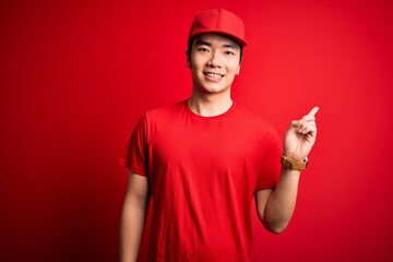 Young handsome chinese delivery man wearing cap standing over isolated red background with a big smile on face, pointing with hand and finger to the side looking at the camera.