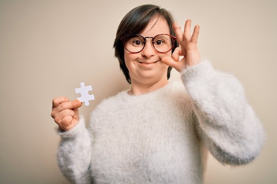 Young down syndrome woman holding puzzle piece as problem solution and cooperation union doing ok sign with fingers, excellent symbol