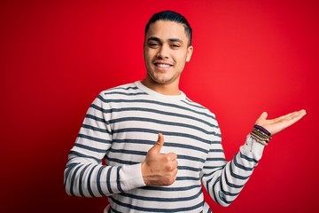 Young brazilian man wearing casual striped t-shirt standing over isolated red background Showing palm hand and doing ok gesture with thumbs up, smiling happy and cheerful