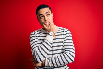 Fototapeta premium Young brazilian man wearing casual striped t-shirt standing over isolated red background Thinking worried about a question, concerned and nervous with hand on chin