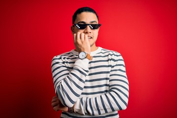 Young brazilian man wearing funny thug life sunglasses over isolated red background looking stressed and nervous with hands on mouth biting nails. Anxiety problem.