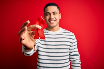 Young brazilian man holding red hiv ribbon standing over isolated background with a happy face...
