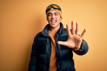 Young brazilian skier man wearing snow sportswear and ski goggles over yellow background showing and pointing up with fingers number five while smiling confident and happy.