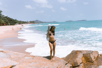 A beautiful girl in a swimsuit is standing on the beach on the rocks with the sea.Against her background,waves and splashes splash.The concept of a traveler and a good life in Thailand.Model