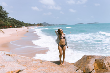 A beautiful girl in a swimsuit is standing on the beach on the rocks with the sea.Against her background,waves and splashes splash.The concept of a traveler and a good life in Thailand.Model