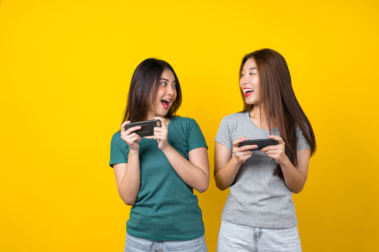 Two Happiness Asian smiling young woman gamer using smart mobile phone and playing games on isolated yellow color background, Lifestyle and leisure with hobby concept
