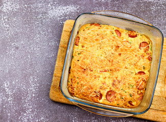 Casserole with cheese, green onions, eggs and sausages in a glass baking dish on a dark background. Top view, flat lay