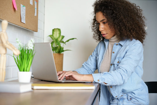 Focused Young African American Teen Girl College Student Using Laptop Computer Typing Studying Working Online. Serious Mixed Race Millennial Woman Doing Internet Research Sitting At Home Office Desk.