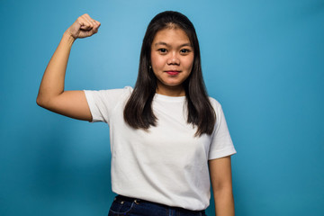 Portrait of Young beautiful asian women using white T-shirt with blue isolated background, raises arms and shows biceps