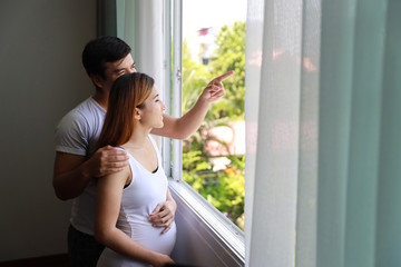 Side view young caucasian husband embracing young pregnant asian belly wife and pointing at something and look away to outside window