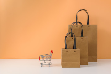Paper shopping bags and shopping cart or trolley on white table and pastel orange background