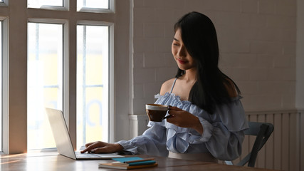 Photo of beautiful woman holding a coffee cup in hand while typing on computer laptop and sitting at the wooden working table over comfortable living room as background.