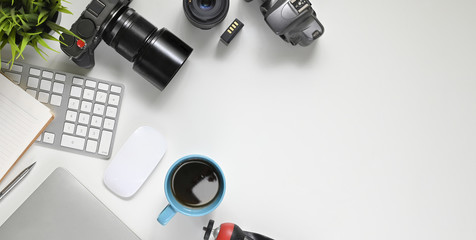 Top view image of photographer working desk with accessories putting on it. Digital camera with lens, wireless keyboard, mouse, coffee cup, note, diary, pen, battery, lens, tripod and potted plant.