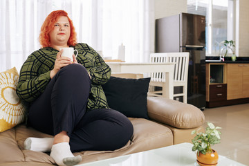 Pensive red haired woman in plaid shirt resting on sofa with big cup of coffee