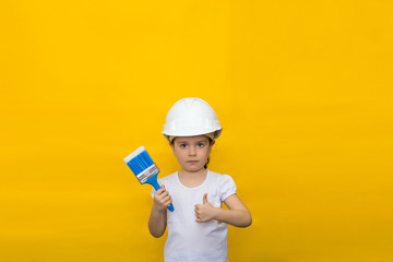 little girl in a construction white helmet holds a paint brush, showing thumb's up gesture on a yellow background