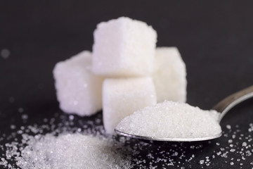 Fototapeta na wymiar Side view of sugar cubes on a gray background and a spoon with sugar crystals. Spoonful of sugar sand close-up on the right side of the frame. Side view, horizontal, free space. Health concept.