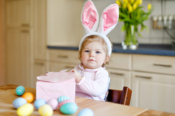 Cute little toddler girl wearing Easter bunny ears playing with colored pastel eggs. Happy baby child unpacking gifts. Adorable healthy smiling kid in pink clothes enjoying family holiday