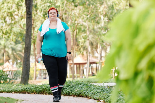 Overweight young woman with towel on her neck walking in park with bottle of fresh water in hand