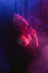 A beautiful slender girl with long hair behind a blind glass in neon lights in a black dress