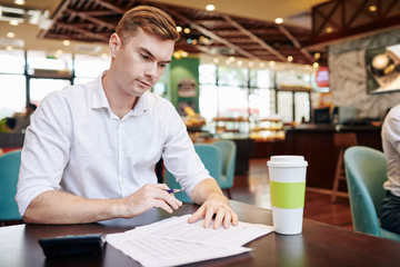 Fototapeta premium Handsome young entrepreneur sitting at cafe table with cup of take away coffee and checking figures in financial report