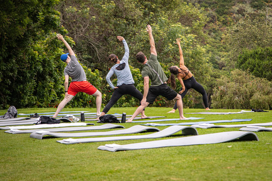 Yoga Instructor And Students Outside On Grass With Yoga Mats Practicing Poses. Trees Are In The Background