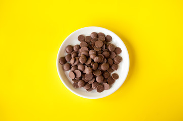 Milk chocolate drops in a plate on a yellow background, top view