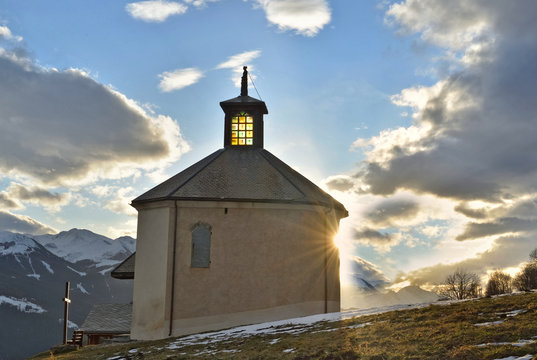 Little Alpine Chapel Lightening By The Sun Under Cloudy Sky At Sunset