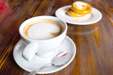 a cup with cappuccino on a saucer is located next to a plate with a bun
