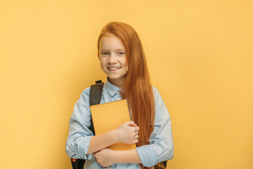 caucasian red haired school girl isolated over yellow background. sweet little girl hug book, stand with bag on her back and smile at camera. school, education concept