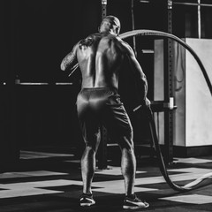 Fototapeta na wymiar Back view of fit sporty man training with battle rope, dramatic black and white image